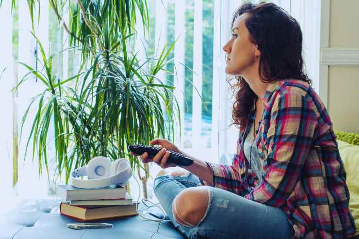 Ragazza seduta in posizione accovacciata su un divano mentre fa zapping con il telecomando della tv