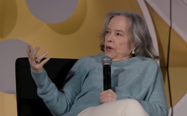 Kathy Bates seduta su una poltrona con il microfono in mano e la mano destra alzata, parla