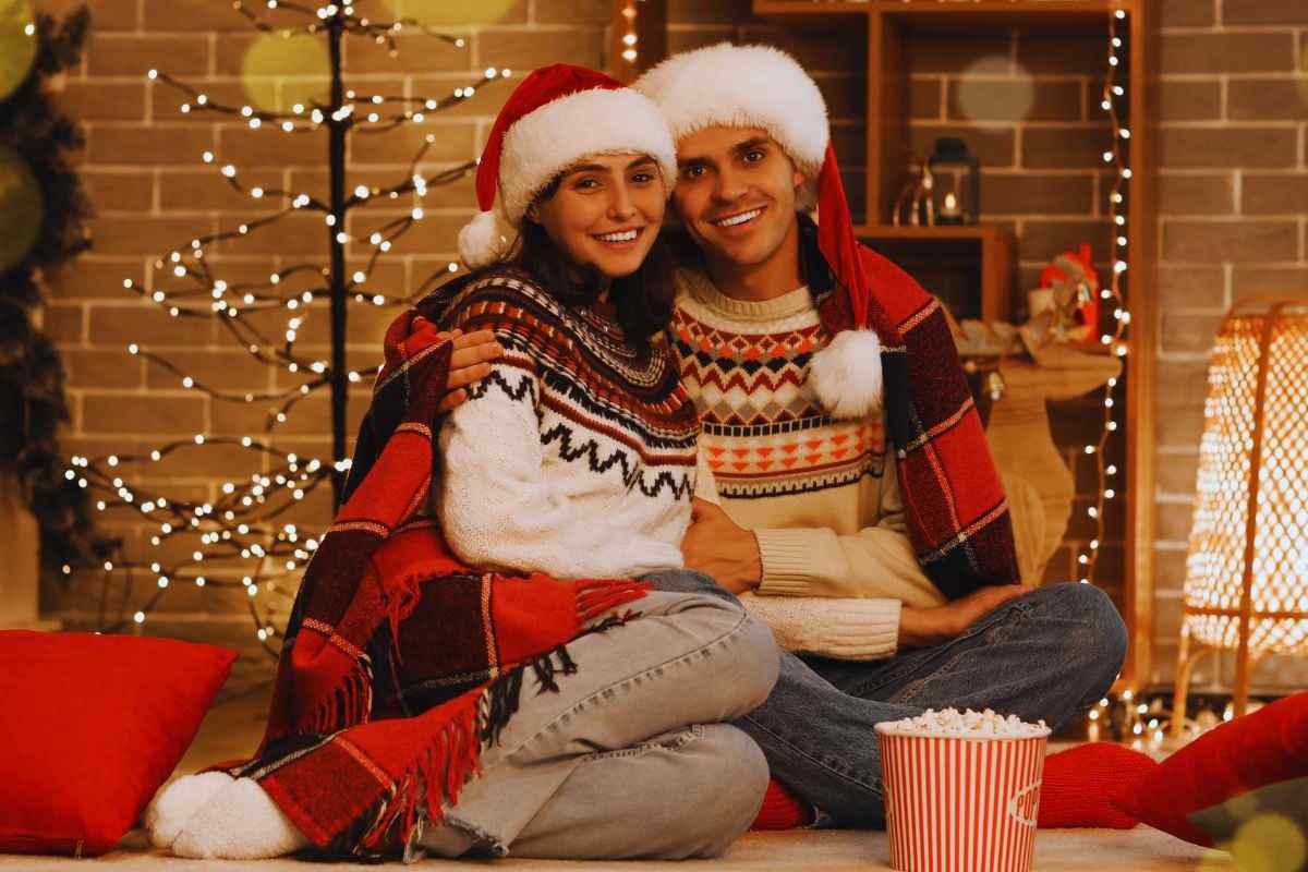 Coppia con indosso i cappelli di Babbo Natale che si stringe sotto la coperta all'interno di una stanza addobbata per le feste e un cestello di popcorn poggiato ai loro piedi