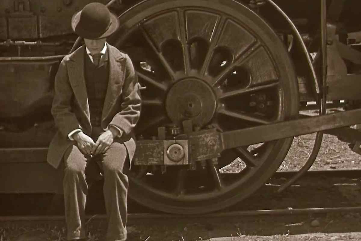 Buster Keaton seduto su uno scalino al suo fianco l'enorme ruota di un treno