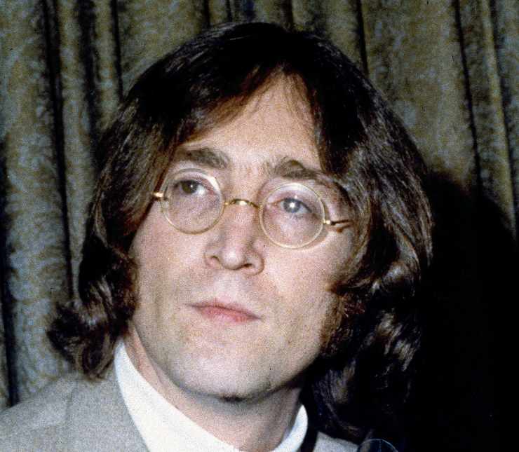 John Lennon guarda a destra con indosso i suoi classici occhialetti tondi e un abito elegante