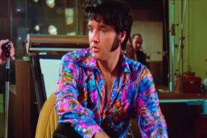 Elvis Presley seduto guarda di lato indossa una camicia molto colorata, lo sguardo è perso nel vuoto