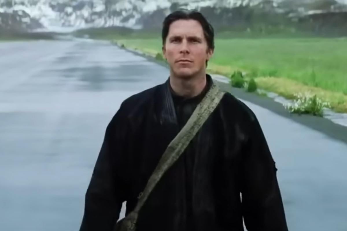 Christian Bale cammina su una strada con del verde intorno