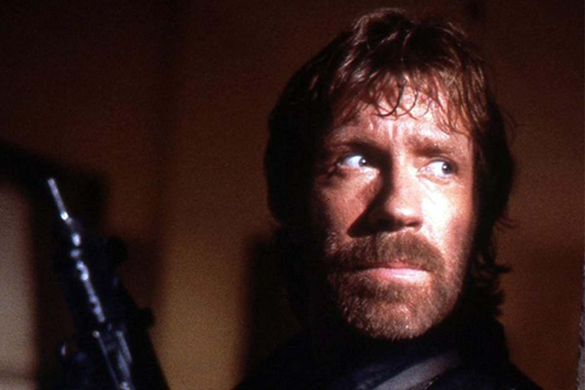 Chuck Norris con in mano un'arma guarda nascosto dietro ad un muro