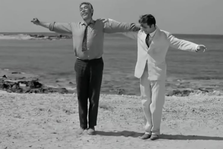 Anthony Quinn che balla con un altro uomo accanto su una spiaggia con il mare sullo sfondo