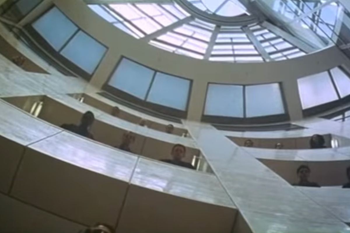 Prospettiva di una stanza vista dal basso che mostra delle finestre in un contesto di cemento con delle persone che si sporgono da un affaccio 